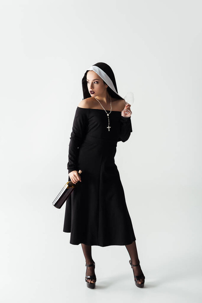 beautiful sexy nun holding glass and bottle of wine on grey - Фото, изображение