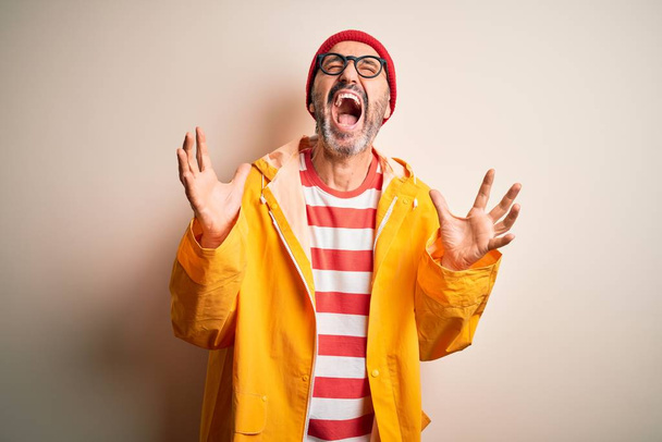 Middle age hoary man wearing glasses and rain coat standing over isolated white background crazy and mad shouting and yelling with aggressive expression and arms raised. Frustration concept. - Photo, Image