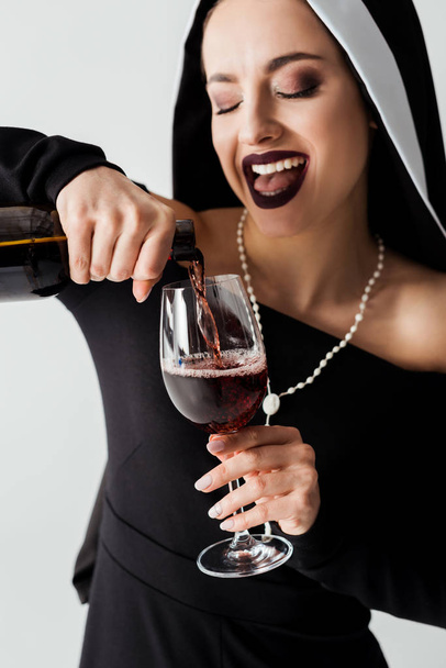 συναισθηματική σέξι καλόγρια ρίχνει κρασί από μπουκάλι σε ποτήρι απομονωμένο σε γκρι - Φωτογραφία, εικόνα