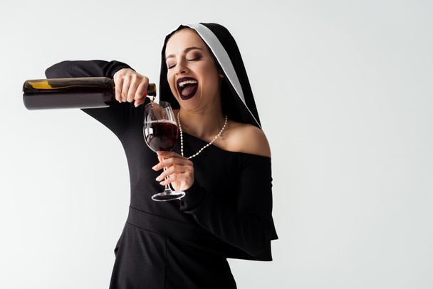 excited sexy nun pouring wine from bottle into glass isolated on grey - Photo, Image
