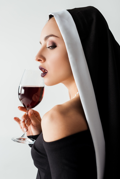 attractive sensual nun holding glass of red wine isolated on grey - Фото, зображення