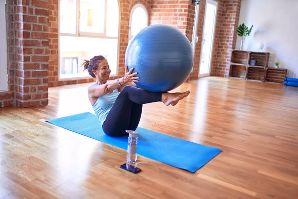 Middle age beautiful sportswoman smiling happy. Doing exercise using fitness ball in a class of yoga at gym - Foto, Imagen