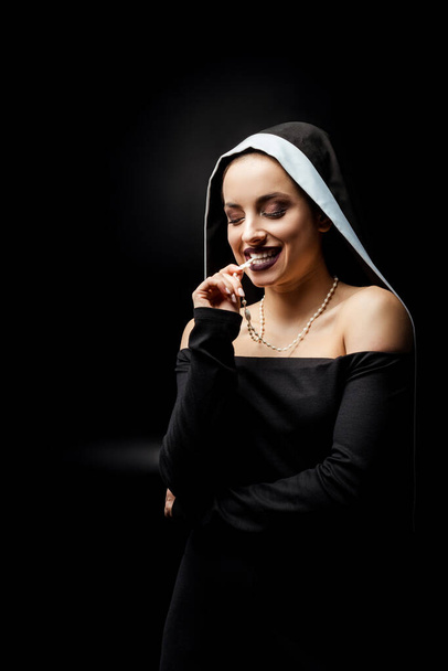 sexy smiling nun in black dress biting cross, isolated on black   - Фото, изображение
