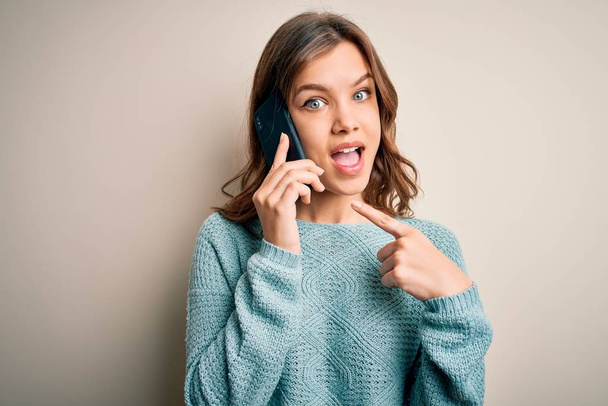 Chica rubia joven teniendo una conversación hablando en el teléfono inteligente sobre un fondo aislado muy feliz señalando con la mano y el dedo
 - Foto, imagen