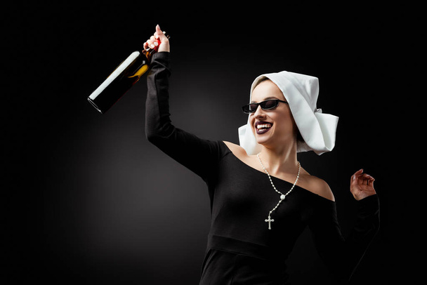 aantrekkelijke lachende non in zonnebril met fles wijn op grijs - Foto, afbeelding