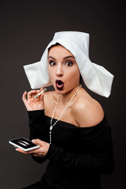 shocked nun holding dollar banknote and smartphone with cocaine lines on grey - Photo, Image