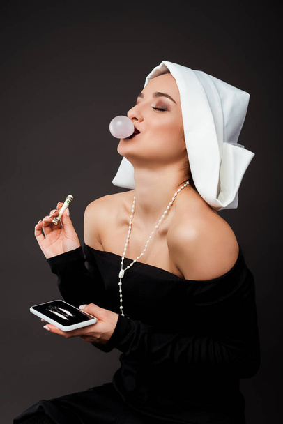 sexy monja riendo con goma de mascar sosteniendo billete de dólar y teléfono inteligente con líneas de cocaína en gris
 - Foto, Imagen