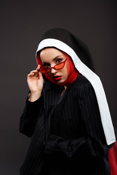 módní sexy démonická jeptiška s červenýma očima pózující v černém obleku, červenými slunečními brýlemi a šátkem, izolovaná na šedé  - Fotografie, Obrázek