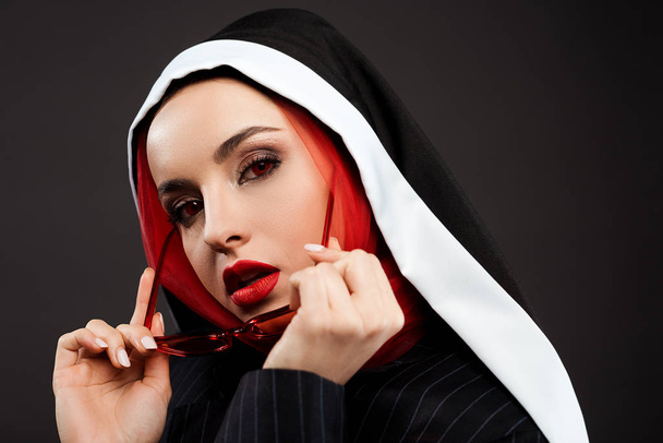 elegante sexy demoníaca freira com olhos vermelhos posando em terno preto, óculos de sol vermelhos e cachecol, isolado em cinza
  - Foto, Imagem