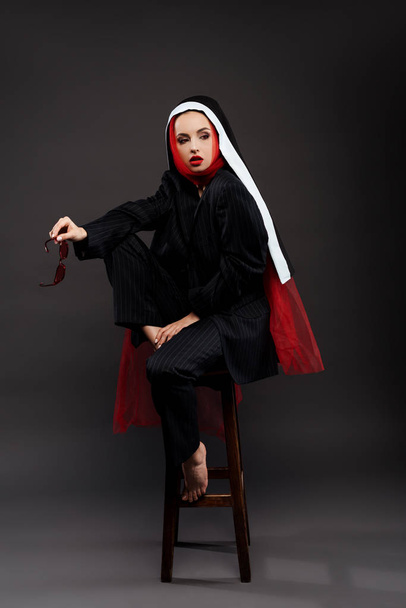 boso stylowe sexy zakonnica pozowanie w czarny garnitur i czerwony szalik w okulary przeciwsłoneczne, na szary  - Zdjęcie, obraz