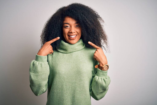 Νεαρή όμορφη Αφροαμερικανή με αφρο μαλλιά που φοράει πράσινο χειμωνιάτικο πουλόβερ χαμογελώντας χαρούμενα δείχνοντας και δείχνοντας με τα δάχτυλα τα δόντια και το στόμα. Έννοια υγείας οδόντων. - Φωτογραφία, εικόνα