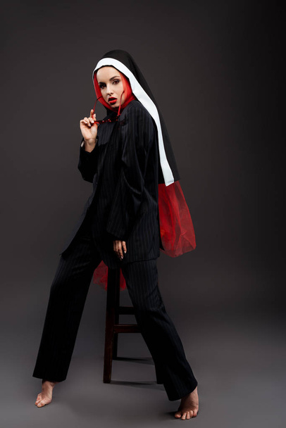 descalça freira sexy na moda posando em terno preto e cachecol vermelho com óculos de sol, em cinza
  - Foto, Imagem