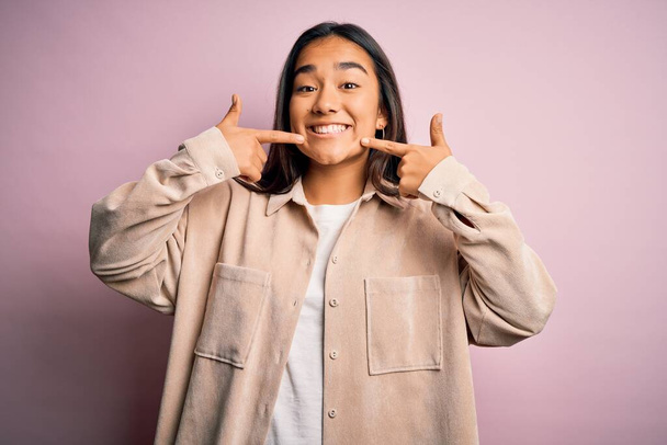 Jeune belle femme asiatique portant chemise décontractée debout sur fond rose souriant montrant joyeuse et pointant avec les doigts dents et la bouche. Concept de santé dentaire
. - Photo, image