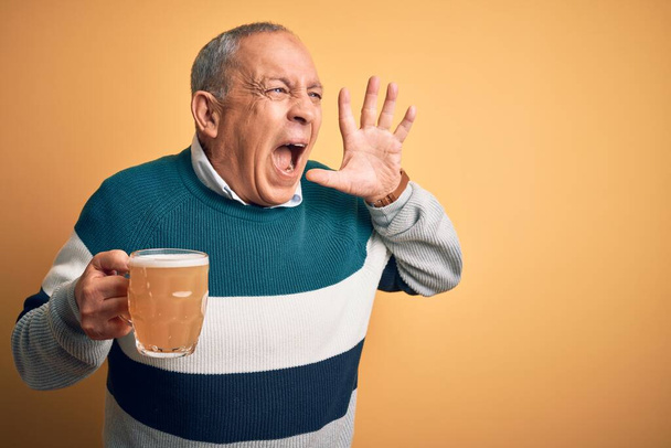 Hoge knappe man die een pot bier drinkt, staand over een geïsoleerde gele achtergrond schreeuwend en schreeuwend met hand op mond. Communicatieconcept. - Foto, afbeelding
