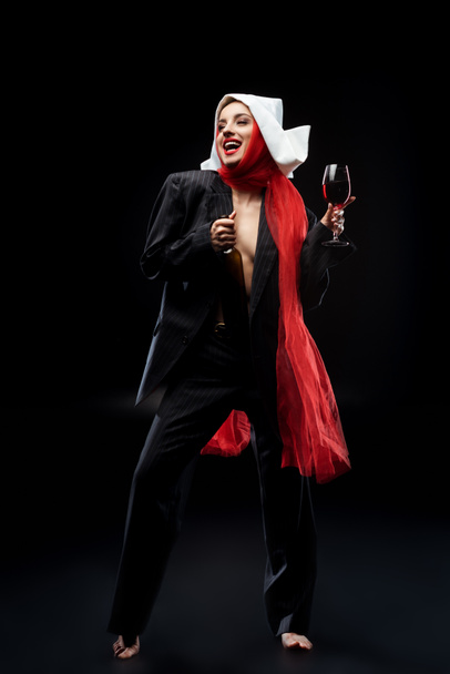 emotional sensual barefoot nun holding glass and bottle of red wine on black - Foto, afbeelding