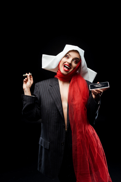 emotional sexy demonic nun holding dollar banknote and smartphone with cocaine lines isolated on black - Photo, Image