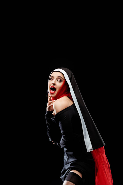 shocked sexy nun posing in black dress, stockings and red scarf, isolated on black - Photo, Image