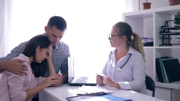 lekarz ze złymi wiadomościami o zdrowiu kobiety, mężczyzna przytula swoją zdenerwowaną żonę siedzącą w biurze w szpitalu - Materiał filmowy, wideo