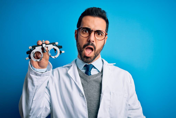 Νεαρός όμορφος οπτικός άντρας με γενειάδα που κρατάει γυαλιά οπτομετρίας πάνω από μπλε φόντο στο σοκαριστικό πρόσωπο, που δείχνει σκεπτικός και σαρκαστικός, έκπληκτος με ανοιχτό στόμα - Φωτογραφία, εικόνα