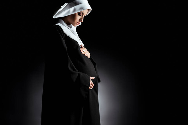 attractive pregnant nun touching tummy on black - Photo, Image