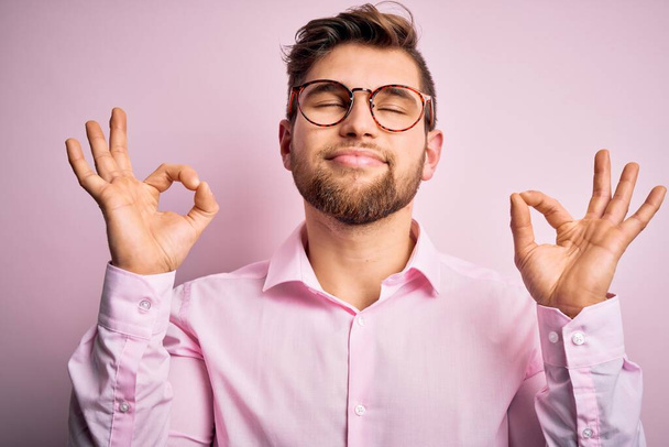 Homem loiro bonito jovem com barba e olhos azuis vestindo camisa rosa e óculos relaxar e sorrir com os olhos fechados fazendo gesto de meditação com os dedos. Conceito de ioga
. - Foto, Imagem