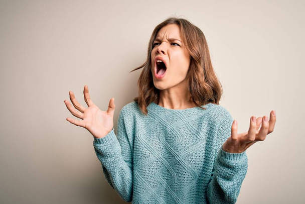 Young blonde girl wearing casual blue winter sweater over isolated background crazy and mad shouting and yelling with aggressive expression and arms raised. Frustration concept. - Photo, Image