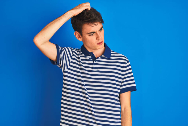 Το έφηβο αγόρι που φοράει casual t-shirt και στέκεται πάνω από το μπλε απομονωμένο φόντο μπερδεύει και αναρωτιέται για την ερώτηση. Αβέβαιο με αμφιβολία, σκέψη με το χέρι στο κεφάλι. Σκεπτική έννοια. - Φωτογραφία, εικόνα