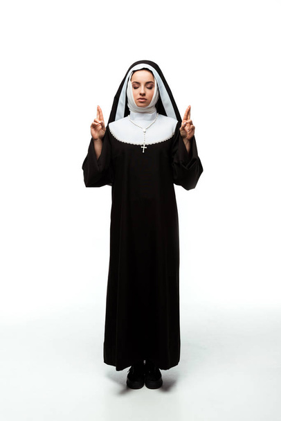 beautiful nun with closed eyes holding fingers crossed, isolated on white - Photo, Image