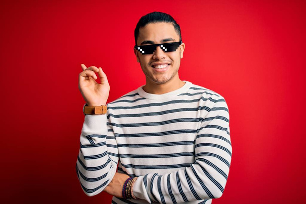 Young brazilian man wearing funny thug life sunglasses over isolated red background with a big smile on face, pointing with hand and finger to the side looking at the camera. - Фото, зображення
