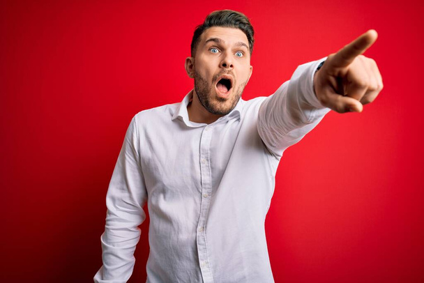 Young business man with blue eyes wearing elegant shirt standing over red isolated background Pointing with finger surprised ahead, open mouth amazed expression, something on the front - Photo, image