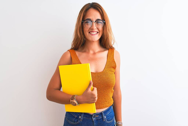 Young beautiful redhead woman wearing glasses reading a book over isolated background with a happy face standing and smiling with a confident smile showing teeth - Photo, Image