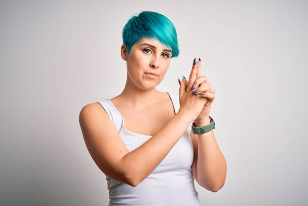 Mujer hermosa joven con el pelo azul de la manera que usa la camiseta casual sobre fondo blanco que sostiene el arma simbólica con gesto de la mano, jugando armas de tiro que matan, cara enojada
 - Foto, Imagen
