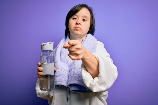 Молодая женщина с синдромом Дауна тренируется в тренажерном зале держа бутылку воды и спортивное полотенце указывая пальцем на камеру и на вас, знак руки, позитивный и уверенный жест спереди
 - Фото, изображение