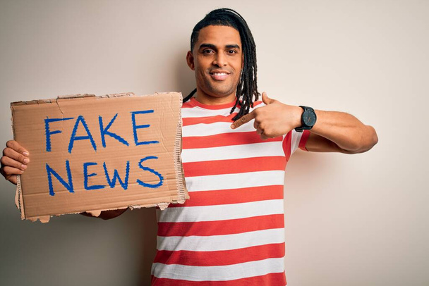 Jeune homme afro-américain avec dreadlocks tenant bannière avec fausses nouvelles message protestation avec visage surprise pointant vers lui-même
 - Photo, image