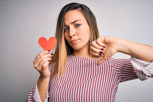 Young beautiful blonde romantic woman holding red paper heart over white background with angry face, negative sign showing dislike with thumbs down, rejection concept - Фото, зображення
