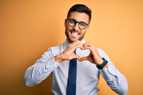 Fiatal jóképű üzletember visel nyakkendő és szemüveg felett áll sárga háttér mosolyog a szerelemben csinál szív szimbólum alakú kezekkel. Romantikus koncepció. - Fotó, kép