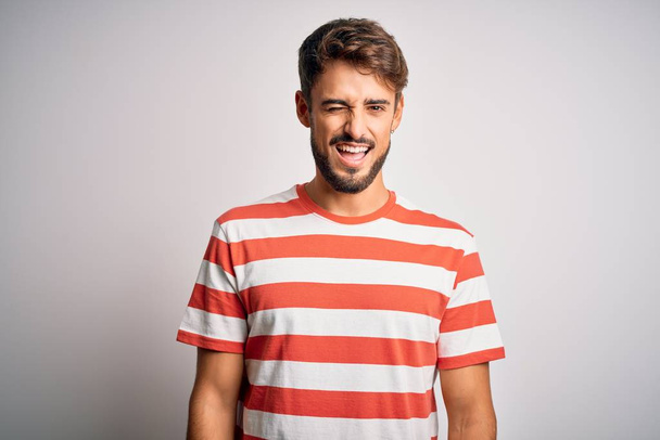 Молодой красивый мужчина с бородой в полосатой футболке стоит на белом фоне подмигивая, глядя в камеру с сексуальным выражением лица, веселым и счастливым лицом
. - Фото, изображение