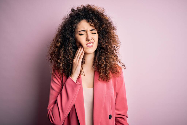 Young beautiful businesswoman with curly hair and piercing wearing elegant jacket touching mouth with hand with painful expression because of toothache or dental illness on teeth. Dentist - Foto, afbeelding