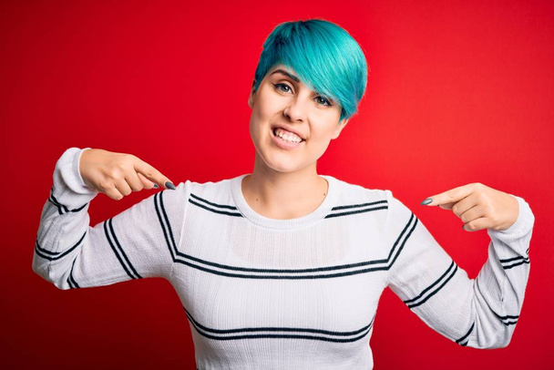 Young beautiful woman with blue fashion hair wearing striped sweater over red background looking confident with smile on face, pointing oneself with fingers proud and happy. - Фото, изображение