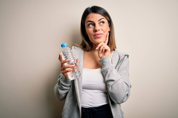 Young beautiful brunette sporty woman drinking bottle of water over isolated white background serious face thinking about question, very confused idea - Fotoğraf, Görsel