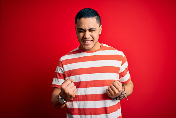 Jonge Braziliaanse man draagt casual gestreept t-shirt staan over geïsoleerde rode achtergrond opgewonden voor succes met opgeheven armen en ogen gesloten vieren overwinning glimlachen. Winnaars concept. - Foto, afbeelding