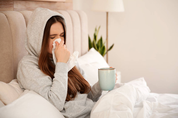 Sick young woman with cup of hot drink in bed at home. Influenza virus - Фото, изображение