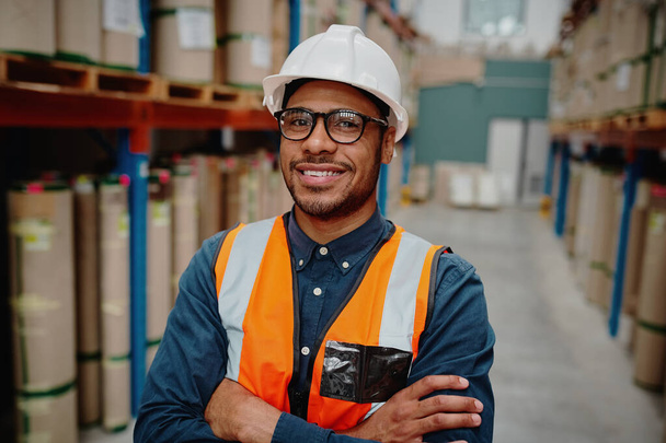 Portrait of handsome cheerful supervisor in warehouse standing with folded hands with material while wearing orange vest and white hardhat - Фото, изображение