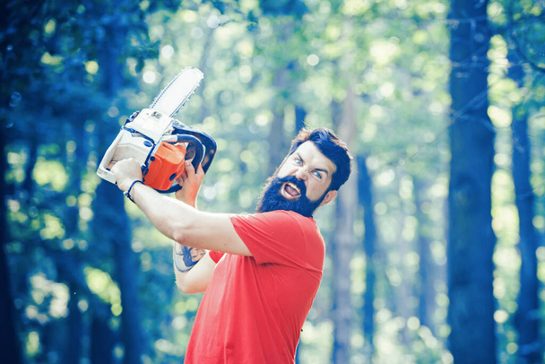 Lumberjack holding the chainsaw. Deforestation is a major cause of land degradation and destabilization of natural ecosystems. Handsome young man with axe near forest. - Foto, Imagen