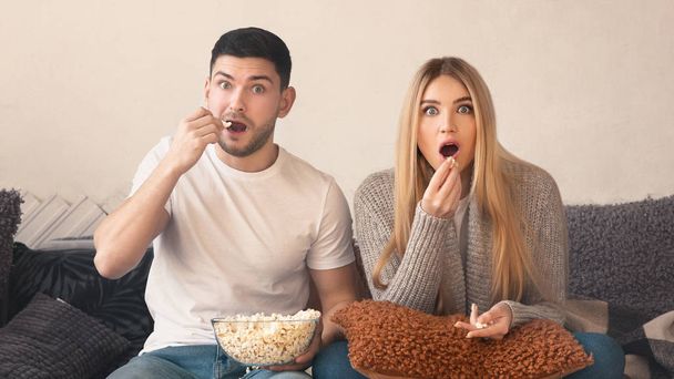 Überraschtes junges Paar mit Popcorn vor dem Fernseher im Wohnzimmer - Foto, Bild
