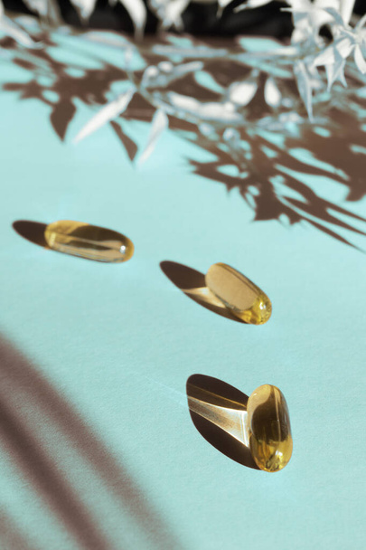 Oil yellow gelatin capsules on blue background with flower shadow, vitamins and antioxidant concept, omega 3 oil for healthcare. Copy space. - Photo, Image