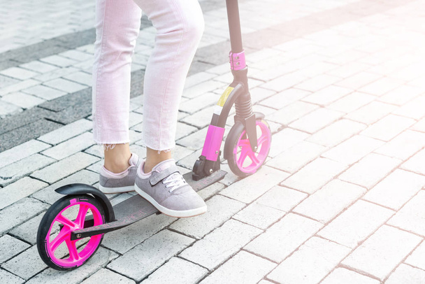 Onderste deel van de jonge volwassen vrouw dragen roze casual jeans en sneakers rijden push kick scooter in de stad straat op zomerse dag buiten. tiener meisje having plezier genieten sport activiteiten buiten. - Foto, afbeelding