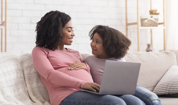 Embarazada mujer negra y poco hija usando portátil juntos en casa
 - Foto, imagen