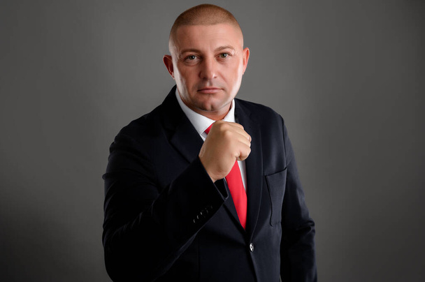 Portret fajnego biznesmena ubranego w elegancki czarny garnitur i czerwony krawat pokazujący pięść odizolowaną od szarego tła - Zdjęcie, obraz