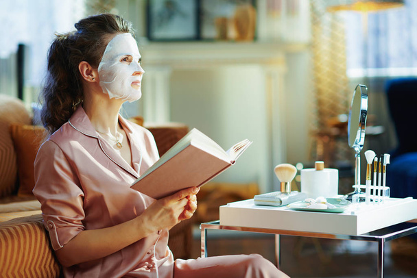 young woman in pajamas with white sheet facial mask on face holding book and looking into the distance in the modern living room in sunny winter day. - Photo, Image